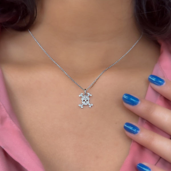 A necklace with a small skull-shaped pendant, pave set with diamonds in a white gold color, worn by a person with blue nails.