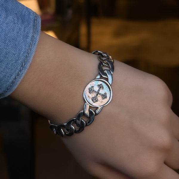 A white gold and silver link bracelet with a black diamond cross seal, worn on a wrist.