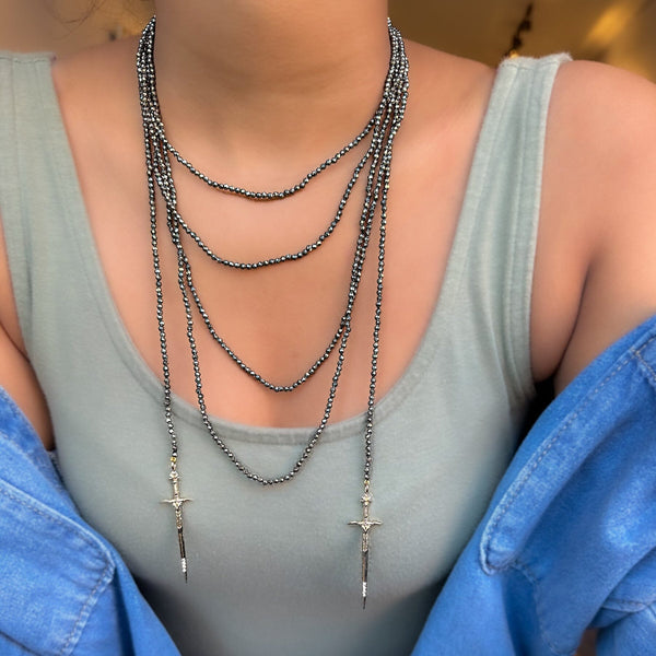 A close-up image of a pyrite beaded necklace with gold diamond daggers, showing a series of round faceted beads and two gold-colored metal pendants.